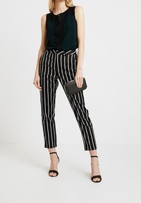 Czarna bezrękawnikowa bluzka z koronkowymi detalami, zestawiona z czarno-białymi spodniami w paski o zwężanym kroju. Modelka trzyma teksturowaną czarną kopertówkę i nosi czarne szpilki.