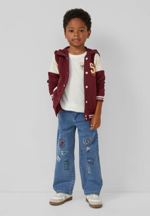 Bordeaux-rode en crème gestreepte varsityjas met knopen over een wit t-shirt; blauwe wijde jeans met verschillende geborduurde patches; witte sneakers.
