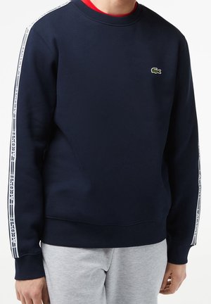 Tmavě modrá mikina s žebrovaným kulatým výstřihem, bílými pruhy po stranách a zeleným logem vyšitým na hrudi. Materiál působí měkce a má texturovaný povrch.