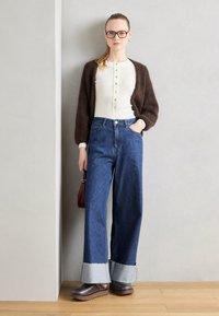 Bruine fluffy cardigan, witte button-up top, hoog getailleerde blauwe denim wijde broek met lichtblauwe manchetten en donkere schoenen, met een rode tas.