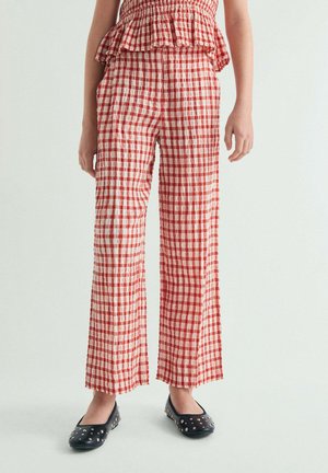 Niño con pantalones sueltos de cuadros rojos y blancos y zapatos planos negros con tachuelas, de pie contra un fondo claro.