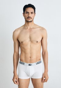 White men's boxer briefs with a black Lacoste logo waistband, fitted design, smooth texture, and mid-length leg. Background is plain.