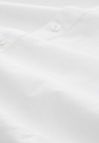 Tessuto di camicia in cotone bianco con una texture morbida, con bottoni visibili e dettagli di cucitura sottili. Il materiale appare leggero e liscio.