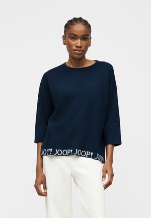 Woman with braided hair wearing a navy blue long-sleeve top with "JOOP!" text on hem and white wide-leg pants against a plain background.