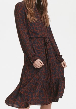 Femme portant une robe à manches longues à imprimé léopard marron et noir, avec des poignets boutonnés et une jupe à volants, tenant légèrement l'ourlet.