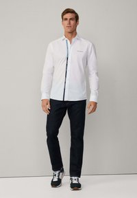 Witte overhemd met contrast blauwe en witte gestreepte knoopsluiting en logo op de borst, gecombineerd met donkere spijkerbroek en sportieve sneakers.