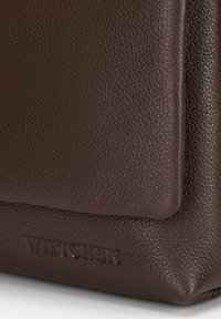 Hnědá kožená taška s texturovaným povrchem, s vyraženým logo. Mezi významné designové prvky patří strukturovaný tvar a hladké okraje.