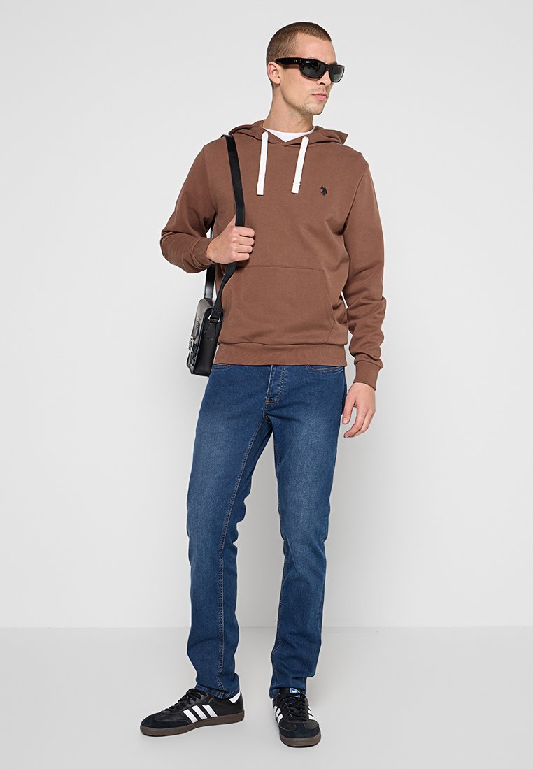 Sudadera marrón con cordones blancos, jeans azul, zapatillas negras y una bolsa cruzada negra; el modelo lleva gafas de sol negras, posando frente a un fondo neutro.