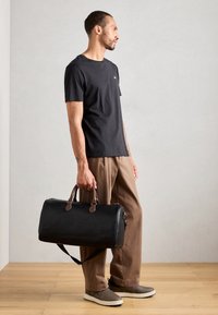 Sac de sport en cuir noir avec des poignées marron, texture lisse, sangle latérale et accessoires métalliques. Porté avec un t-shirt noir et un pantalon beige.