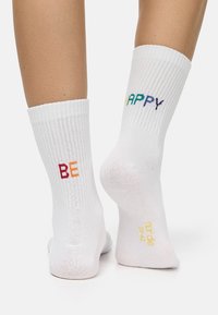 White cotton socks featuring ribbed texture. The words "BE" in gradient warm colors and "HAPPY" in various cool colors are woven on the sides.