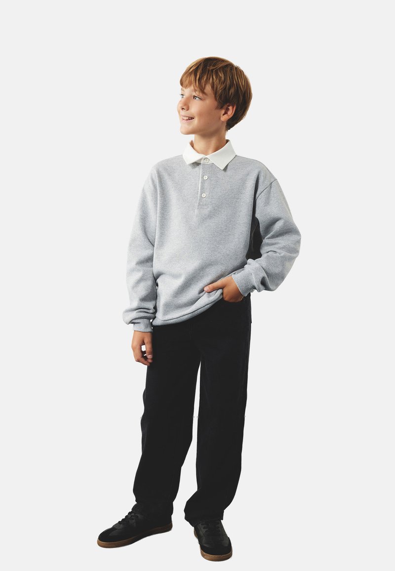 Sudadera gris con cuello blanco, tapeta de tres botones, mangas largas, combinada con pantalones negros y zapatillas negras, en pose de pie.