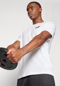 Witte t-shirt met korte mouwen en zwart Puma-logo, vastgehouden door een persoon die een zwart gewichtsschijf optilt, met zichtbare gespierde armen.