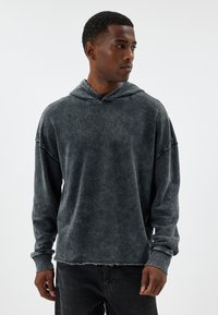 Felpa grigia scura sbiadita con un fit rilassato, maniche lunghe, grande cappuccio e dettagli di orlo grezzo. Tessuto morbido e testurizzato, design minimale.