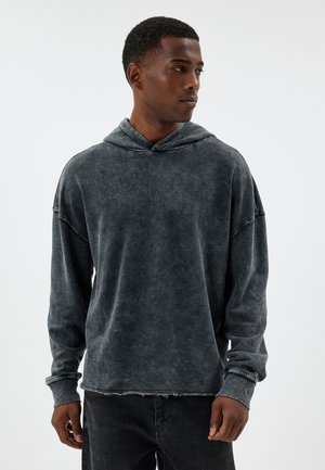 Sudadera desgastada de color gris oscuro con un corte relajado, mangas largas, gran capucha y detalles deshilachados en el dobladillo. Tejido suave y texturizado, diseño minimalista.