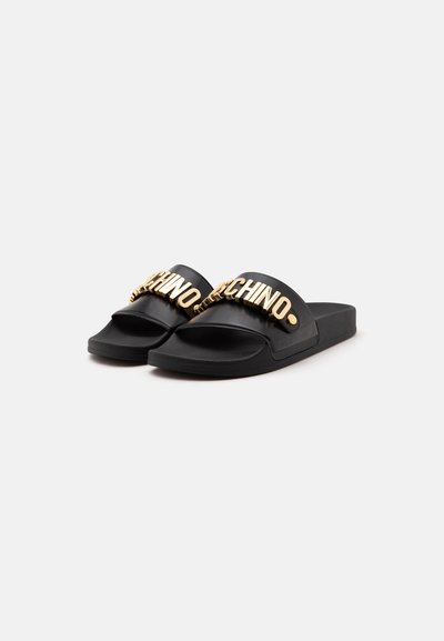 Paire de sandales slides noires avec des lettres dorées en relief formant "MOSCHINO" sur les brides, présentées sur un fond clair uni.