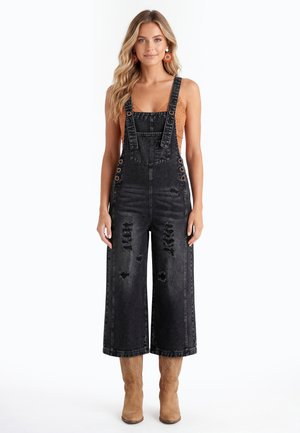Vrouw staat en draagt zwarte versleten denim tuinbroek, bruine gehaakte top eronder, beige laarzen en grote oranje oorbellen tegen een witte achtergrond.