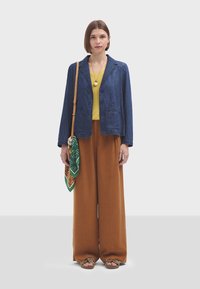 Veste en denim avec des boutons violets, haut jaune, pantalon large marron et écharpe verte à motifs. Matériaux texturés avec des teintes variées.