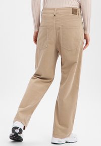 Beige wide-leg pants with a soft texture, featuring two back pockets and a branded tag at the waistband, paired with white sneakers.