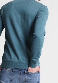 Felpa teal a maniche lunghe con scollatura rotonda e polsini a coste. Presenta una texture morbida e una vestibilità rilassata, abbinata a jeans in denim blu.