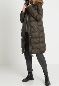 Cappotto imbottito verde oliva con cappuccio in pelliccia sintetica, tasche laterali con zip e texture trapuntata. Indossato con pantaloni grigi e stivali neri lucidi alla caviglia.