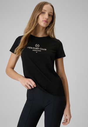 Młoda kobieta o długich blond włosach nosi czarną koszulkę "Victory Club" i czarne legginsy, pozując na szarym tle.