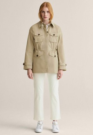 Kvinde står iført beige knappet jakke med fire klaplommer, hvide bukser og hvide sneakers på neutral baggrund.