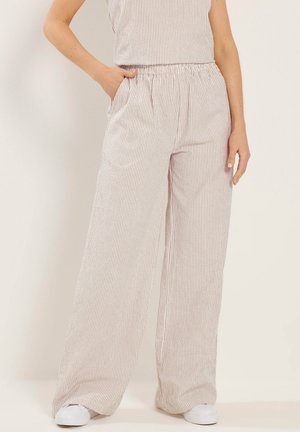 Kobieta nosząca spodnie z wysokim stanem i szerokimi nogawkami w pionowe beżowo-białe paski, w białych trampkach, z ręką w kieszeni.