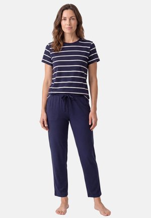 Femme debout pieds nus portant une chemise à manches courtes rayée bleu marine et blanche avec un pantalon bleu marine à cordon, sur un fond uni.