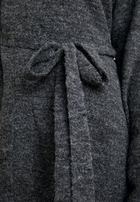 Närbild av en mörkgrå ullkappa med ett knytbälte i midjan, som visar mjuk, strukturerad tyg och mysiga stickade detaljer.