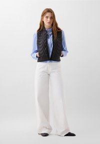 Gilet trapuntato nero con motivo testurizzato, indossato sopra una camicia a righe blu e bianche, abbinato a pantaloni bianchi a gamba larga e scarpe nere.