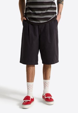 Persoon draagt zwarte knielange shorts, witte geruite sokken, rode sneakers en een zwart-wit gestreept shirt met handen in de zakken.