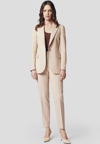 Kremowy, dopasowany garnitur z gładkiego materiału, z marynarką na jeden guzik i wąskimi spodniami. Akcentowany minimalnymi detalami.