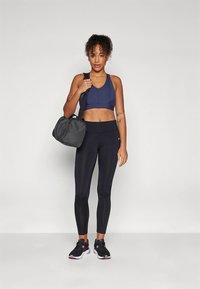 Granatowy sportowy stanik, czarne legginsy, czarne buty sportowe z różowymi akcentami oraz szara torba na siłownię. Modelka stoi prosto, zwrócona przodem, z neutralnym wyrazem twarzy.