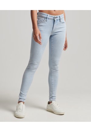 Jean slim fit bleu clair porté avec des baskets blanches, montré du milieu du torse aux pieds sur un fond neutre uni.