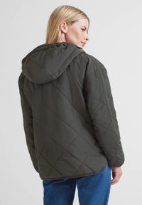 Woman wearing a hooded, dark green quilted jacket and blue jeans, facing away with a side profile visible against a plain background.