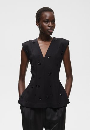 Femme à la peau foncée portant un haut noir sans manches avec un profond décolleté en V et de petites ornements décoratifs, associé à un pantalon noir ample.