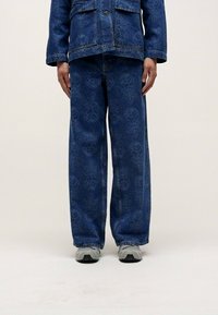 Blaue Jeansjacke und weite Hosen mit einem dezenten Blumenmuster. Das Outfit wird mit grauen Sneakers auf einem neutralen Hintergrund kombiniert.