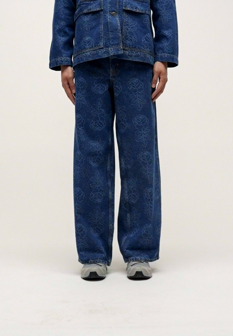 Veste en denim bleu et pantalon large présentant un motif floral discret. L'ensemble est associé à des baskets grises sur un fond neutre.