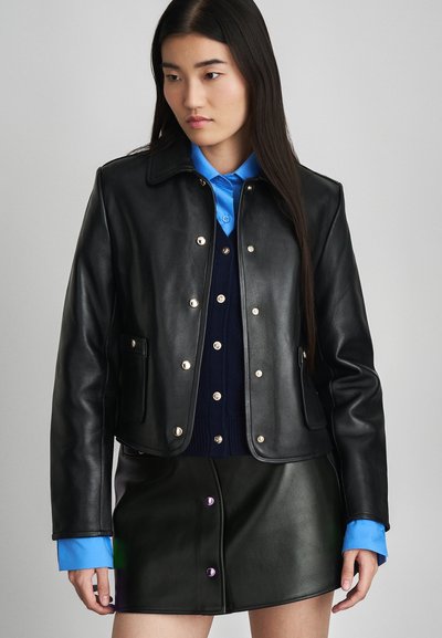 Veste en cuir noire avec un col, du matériel doré, associée à une chemise bleue à col et une jupe en cuir verte avec des détails de boutons.