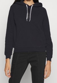 Marinblå hoodie med framficka, vita dragband och broderad design på bröstet. Har ribbade ärmslut och nederkant.