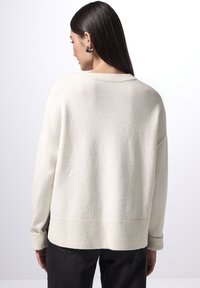 Jasna kremowa, oversize sweter z ribbowanymi mankietami i dołem, wykonany z teksturowanej tkaniny. Model nosi czarne dżinsy, odwracając się od kamery.
