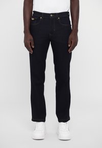 Σκούρο μπλε τζιν παντελόνι με ίσια γραμμή, που διαθέτει κίτρινες ραφές και κλείσιμο με κουμπί μπροστά. Συνδυάζεται με λευκά αθλητικά παπούτσια.