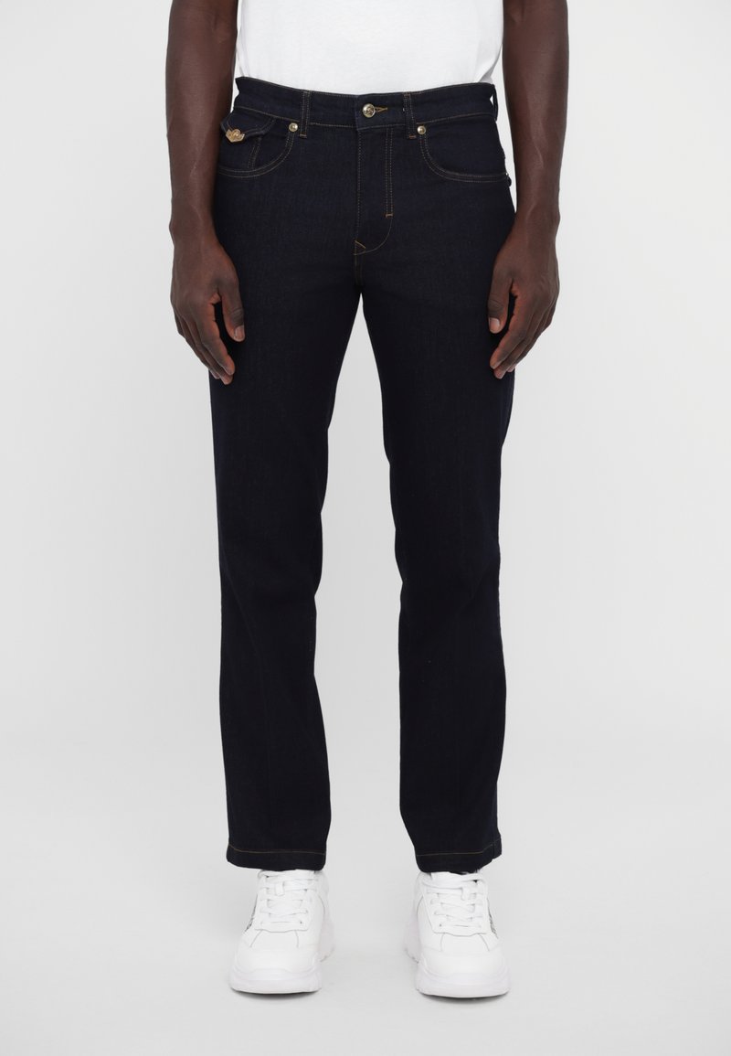 Σκούρο μπλε τζιν παντελόνι με ίσια γραμμή, που διαθέτει κίτρινες ραφές και κλείσιμο με κουμπί μπροστά. Συνδυάζεται με λευκά αθλητικά παπούτσια.