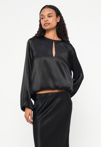 Top din satin negru cu decolteu în forma de ochi de cheie, mâneci lungi cu volum și croi scurt, asortat cu o fustă din satin negru similară. Textură netedă.