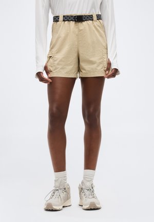 Άτομο που φοράει μπεζ cargo σορτς με μαύρη ζώνη με σχέδια, λευκή μακρυμάνικη μπλούζα, λευκές κάλτσες και μπεζ πεζοπορικά παπούτσια.