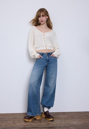 Mlada ženska z ohlapnimi kavbojkami, belim krajšim kardiganom, lesenimi klompi in velikimi uhani stoji ob belem zidu z rokami v žepih.