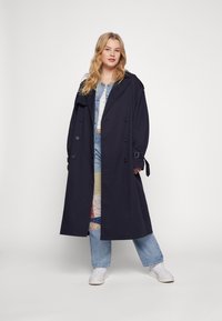 Cappotto navy con design doppiopetto, ampio colletto e maniche con cintura. Indossato sopra un insieme di denim, che presenta una gonna patchwork.