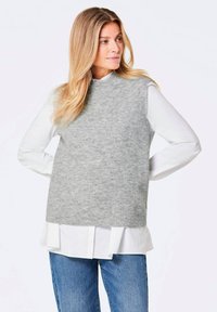 Un gilet en maille grise avec un col montant présentant une texture côtelée, superposé sur une chemise blanche à ourlet plissé, associé à un jean bleu.