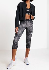 Czarna krótka kurtka, czysty stanik sportowy z białym logo, leggingsy w szarym i czarnym wzorze oraz białe sneakersy z czerwonymi i niebieskimi akcentami.