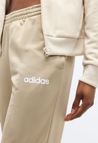 Κοντινό πλάνο ενός ατόμου που φοράει μπεζ φόρμα Adidas και το αντίστοιχο ζακέτα με φερμουάρ, με το λογότυπο της μάρκας να είναι ορατό στο παντελόνι.